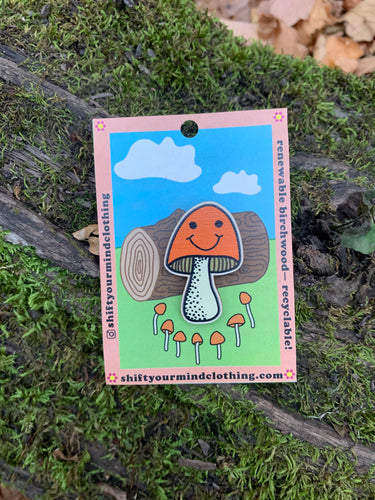 Orange smiling mushroom pin on cardstock packaging featuring a log and other little mushrooms. Pin is sitting on a mossy log in the forest in the photo of the product.